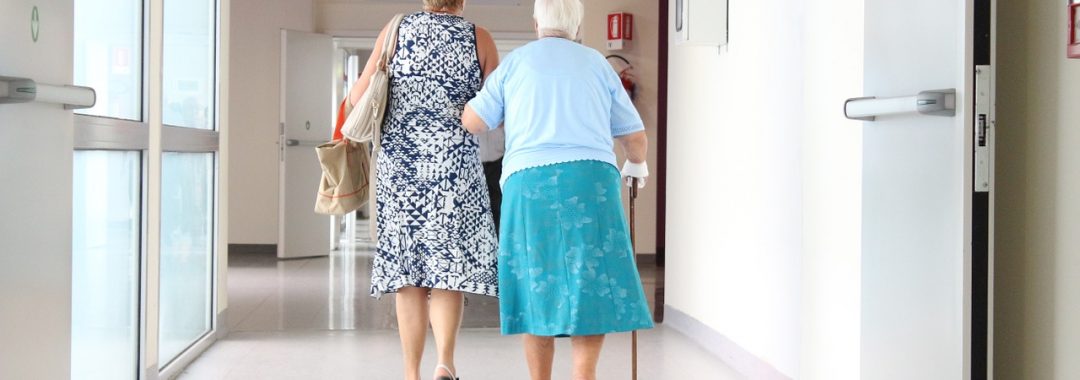 an elderly mother with a walking stick holding onto her daughter whilst walking through a hospital